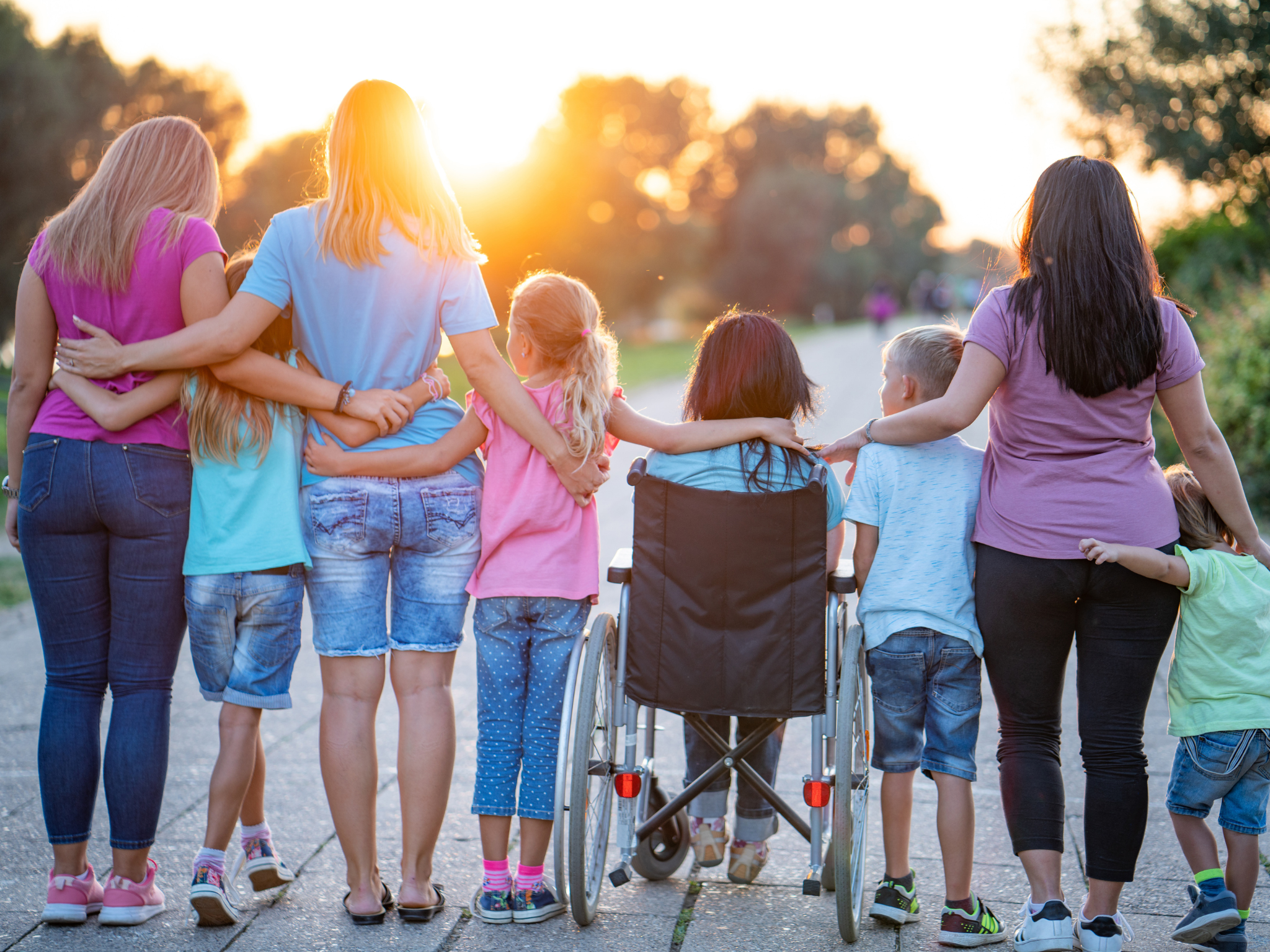 An inclusive family outing in the park, with a child in a wheelchair joining the fun. They all have their arms wrapped around one another and their backs to the camera. The sun is shining through the trees off in the distance.